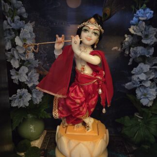 Marble deity of Krishna standing in a lotus base. Dressed in red outfit against a dark background