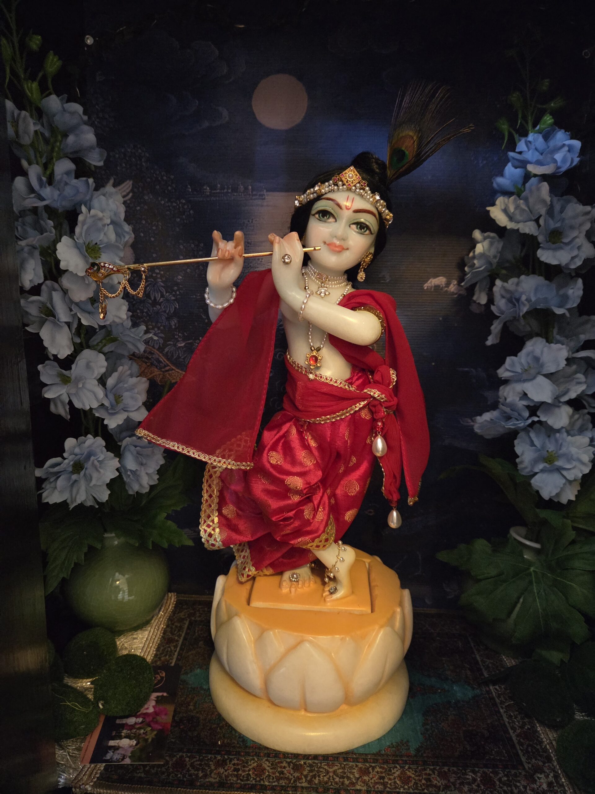 Marble deity of Krishna standing in a lotus base. Dressed in red outfit against a dark background