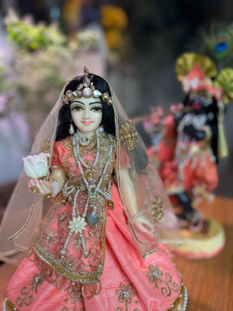 Radha deity carved in marble with a subtle yellow hue dressed in peach shringar stands with Krishna deity behind, carved in black marble, both deities on special cement bases